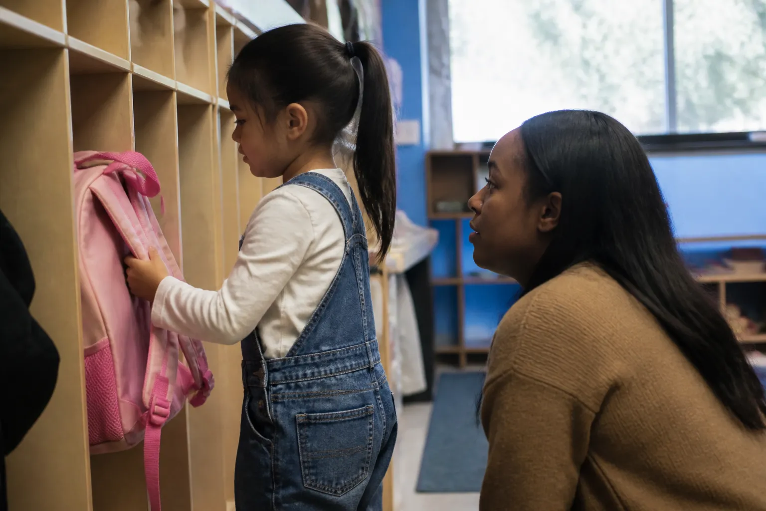 A Friend kneels at eye level beside a young child hanging a backpack in a Houston classroom cubby.