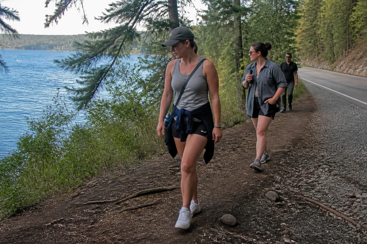 Walkers pass a worn section of the English Point Trail near Hayden Lake Road with pines and blue water behind.